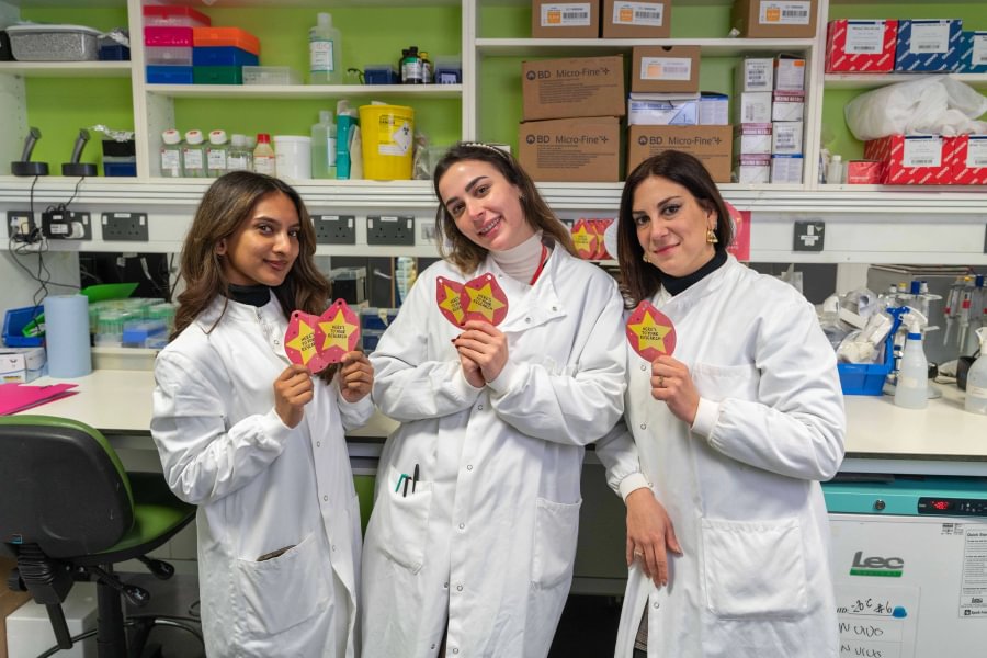 Researchers Priya, Gheed and Roberta in white lab coats, posing for a photo in the lab, each holding a Christmas Star note.