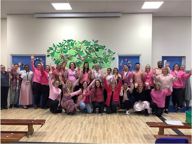 A big group of people in a school hall, wearing pink clothes and waving at the camera