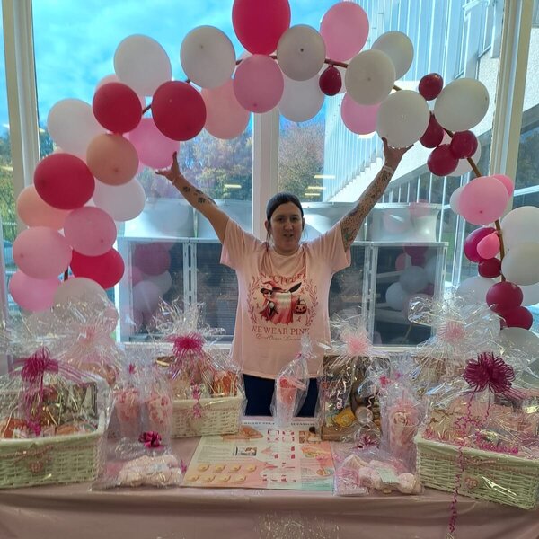 A photo of a woman wearing pink standing behind a table full of gift-wrapped hampers. She is raising her hands to a pink balloon arch overhead