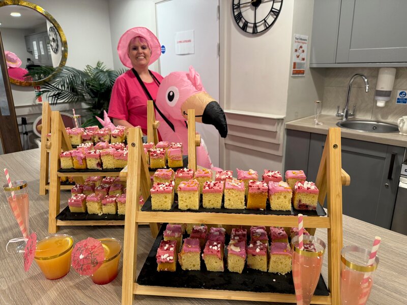 A photo of a woman wearing a pink flamingo inflatable and a pink T-shirt standing behind a table with cakes with pink icing.