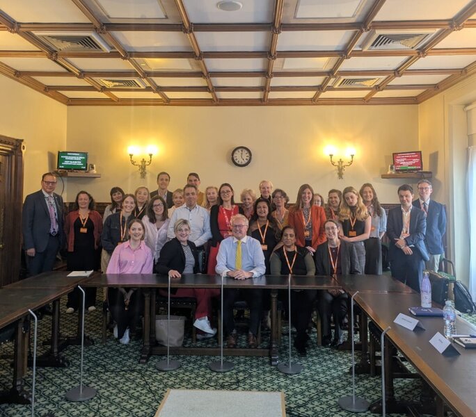 A group of 28 adults sitting and standing round a table smiling at the camera. They are dressed smartly.
