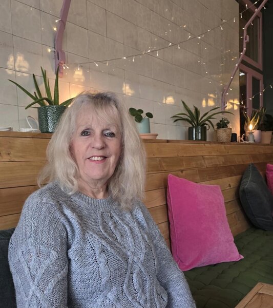 A woman wearing a light grey knitted jumper, sitting on a bench inside a cafe. There are plants and fairy lights in the background. She is smiling at the camera.
