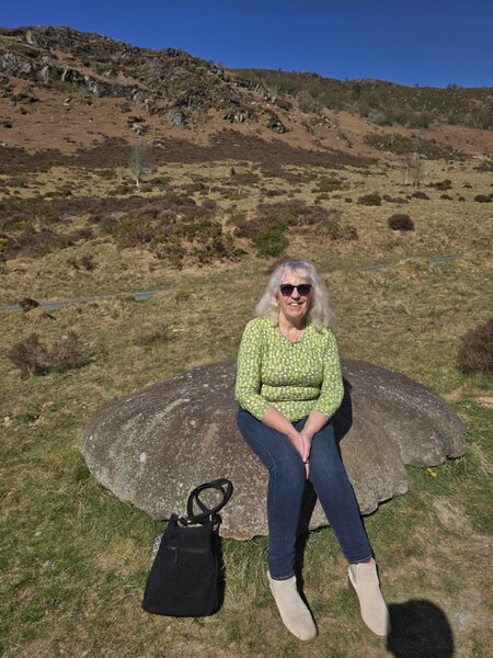 A woman wearing a green top sitting on a rock on a hillside. Behind her there are trees and shrubs and a clear blue sky.