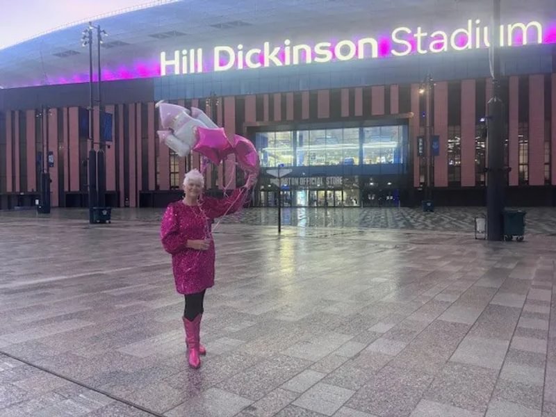 A photo of Julie wearing a pink sparkly dress and pink boots, holding pink and white helium balloons in front of the Hill Dickinson Stadium, the sign for which is lit up pink.