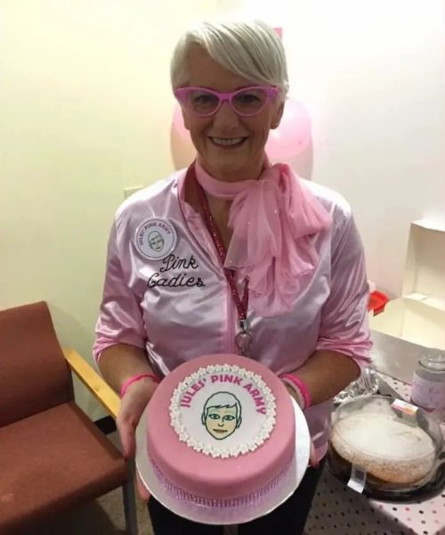 A photo of Julie wearing a pink jacket and pink glasses. She has short hair and is smiling. She is holding a circular pink cake which says "Jules' Pink Army" in the icing.