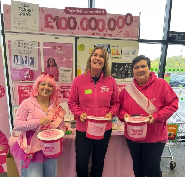 A photo of 3 women taking part in an Asda Tickled Pink bucket collection in an Asda store. They are wearing pink and holding collection buckets in front of a pink stall with a sign that says "Together we've raised £100000000 and counting..."