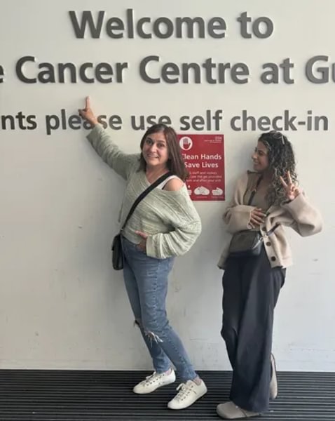 A photo of Evie and her mum standing together in a front of a white wall. They are both smiling and Evie's mum is pointing at a sign that says "Welcome to the Cancer Centre"
