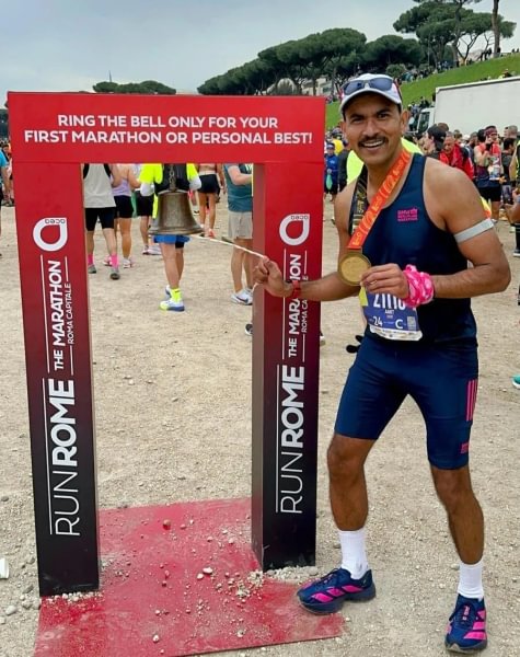 A photo of Amit wearing running kit and a medal at the Rome marathon, ringing a bell for getting a personal best.