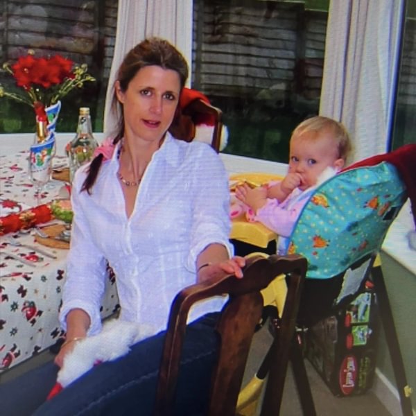 Photo of Sarah as a toddler in a high chair sitting next to her mum at a Christmas dinner.
