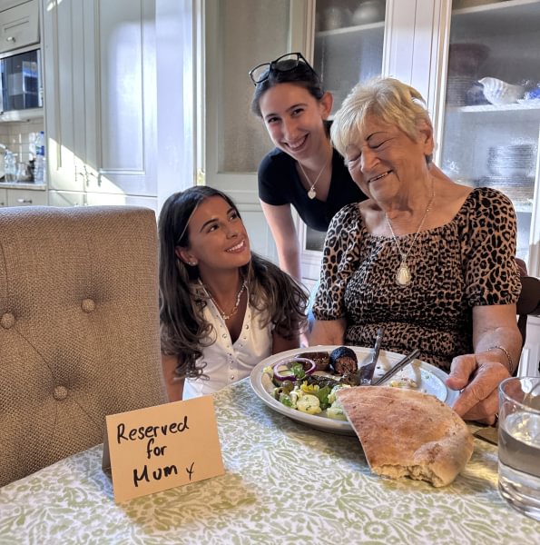 A photo of Evie, her sister and an older relative at a dinner table, smiling. Next to them is an empty place setting with a card that says "Reserved for Mum x".