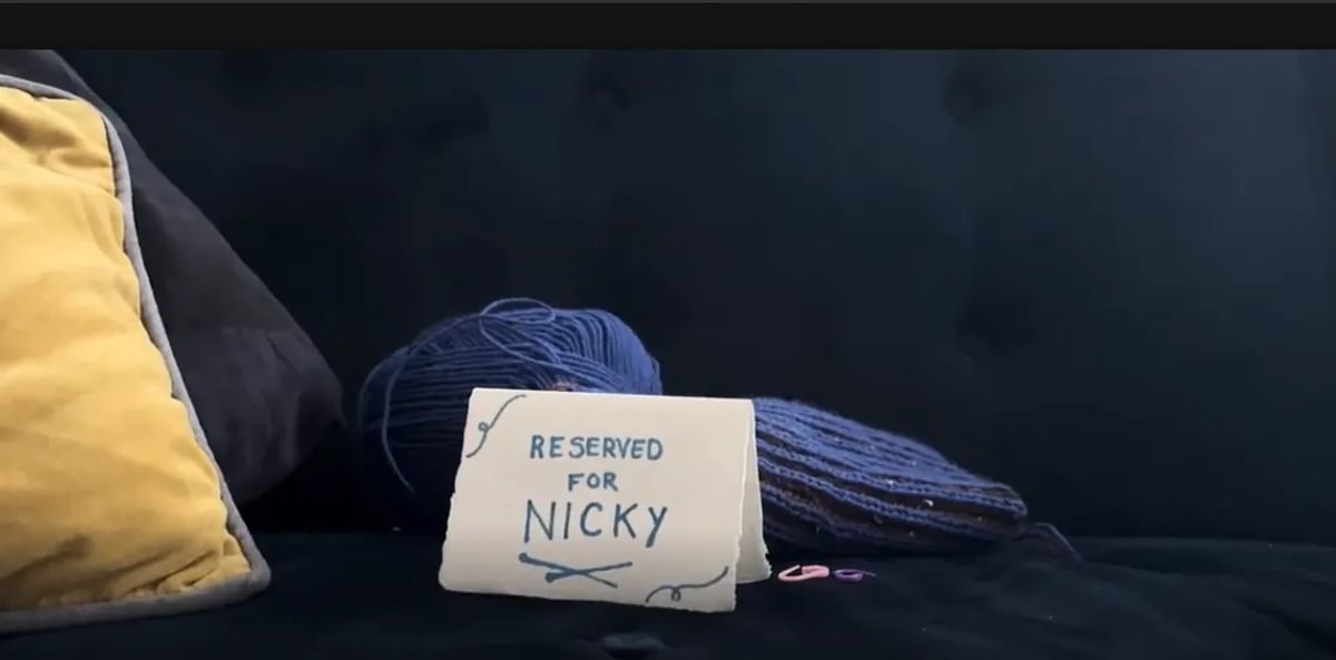 A photo of a handwritten card on a sofa that reads "Reserved for Nicky". Behind it are some cushions and some knitting.
