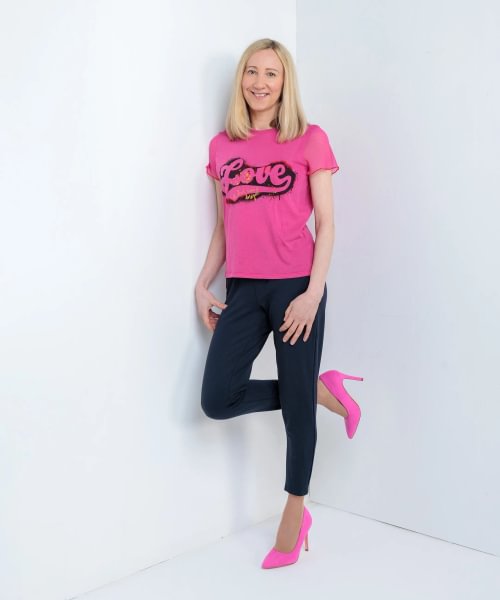 Christina has blonde hair. She's wearing a pink T-shirt with the word 'Love' on it with black leggings and pink high heels.