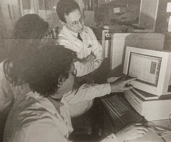 A black and white newspaper photo of Professor Mike Stratton and 2 other research colleagues gathered around a computer. They are all wearing lab coats and one is pointing to a graph on the computer monitor.
