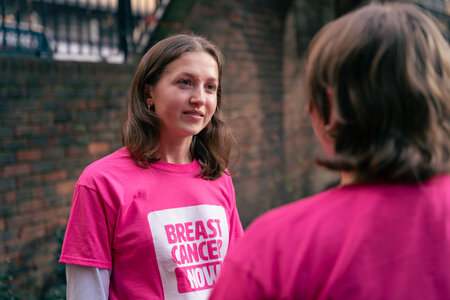 Breast Cancer Now staff wearing merchandise. Two young women talking outside, wearing bright pink Breast Cancer Now cotton t-shirts.