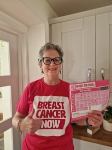 Kerry is standing inside her house wearing her Breast Cancer Now t-shirt and holding her Walk 100 Miles distance tracker.