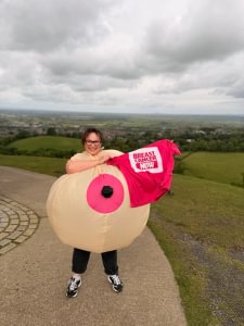 Rachael is wearing an inflatable breast costume and holding her pink Breast Cancer Now t-shirt. She's standing outside with countryside behind her and smiling at the camera.