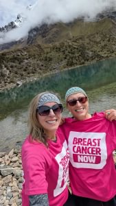 A selfie of Emma and her friend by a small lake surrounded by a rocky landscape. They're both wearing Breast Cancer Now t-shirts and smiling at the camera.