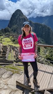 Emma is standing at Machu Picchu in Peru wearing her Breast Cancer Now t-shirt and holding her Walk 100 Miles distance tracker. She's smiling at the camera.