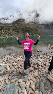 A woman wearing a Breast Cancer Now t-shirt is standing in front of a small lake surrounded by rocky landscape. She's holding her arms up and out to the side and is smiling at the camera.