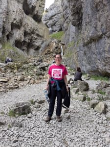 Sarah is standing in front of impressive rocky cliffs, smiling and wearing a pink t-shirt with the Breast Cancer Now logo on it.