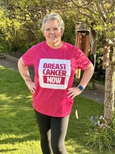 A woman standing in a garden with trees and flowers behind her. She is proudly wearing a pink T-shirt with the Breast Cancer Now logo.