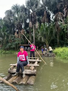Emma and Jo sitting on a raft made out of logs. They're wearing their pink Breast Cancer Now t-shirts and one of them is steering the raft using a very long branch.