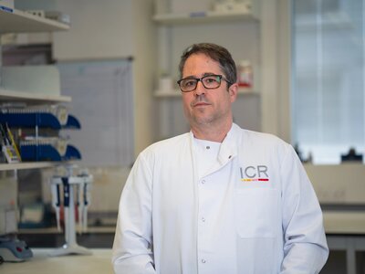 Issac, a man with short grey hair, black square frame glasses, in a white BCN lab coat, posing for portraits in a white lab coat with the lab in the background.