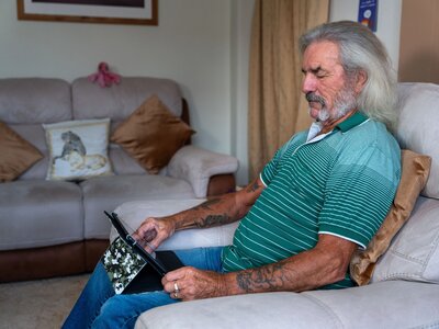 Tony, a man with shoulder length white hair, dark eyebrows, a gold earring and a handlebar moustache, wearing a green and white striped polo t-shirt, with some tattoos visible on his arms. Sat on a cream armchair, using/looking at his tablet, as he would when using the VMU.