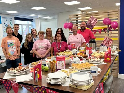 A group of men and women stood behind a table with a spread of food, including scones, cupcakes and apples. The room is decorated with Afternoon Tea bunting and pink balloons.