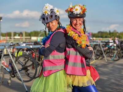 A photo of 2 women who participated in the Women V Cancer Ride the Night cycle event. They are standing back to back with arms folded, smiling, wearing bike helmets decorated with flowers, fluorescent tabbards and tutus over cycling gear. In the background is a bike park.