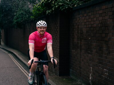 Breast Cancer Now staff wearing merchandise. Staff member Simon, wearing Breast Cancer Now cycling jersey, whilst outside on his bike.