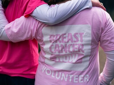 Two people wearing pink cotton t-shirts, with arms around eachother. The words "volunteer" on the back of the top and Breast Cancer Now logo.