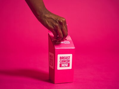Breast Cancer Now paper pop-up collection donation box on a plain pink backdrop, with a hand putting money in the box.