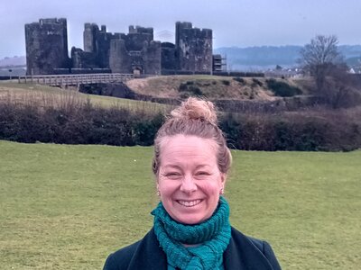 A photo of Sandra smiling at the camera. She is wearing a blue coat and scarf, and standing in front of a castle in England.
