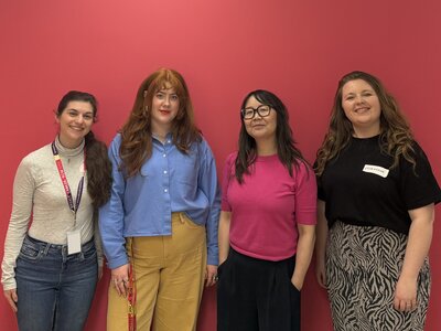 A group of 4 women standing in front of a pink wall. They're all smiling at the camera.