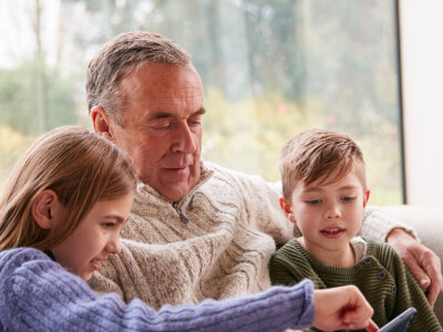 An older man sitting with two young children on a sofa. They're all looking at something on a mobile phone.