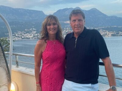 A photo of a couple standing on a balcony on holiday with a view of the ocean and mountains behind them. The woman is wearing a pink dress and the man is wearing pale blue shorts with a navy blue shirt.