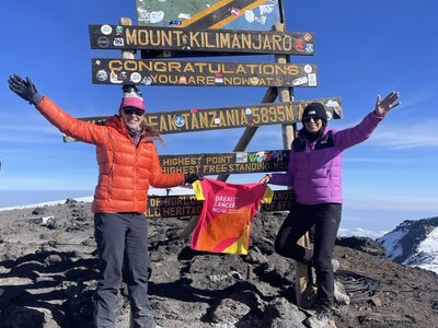 A photo of two trekkers at Mount Kilimanjaro, posing next to a sign at the summit with a Breast Cancer Now t-shirt.