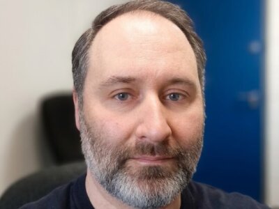 Portrait photo of Christopher Staples, who has blue eyes, grey-brown hair and a beard.