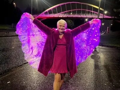 A photo of Julie wearing a pink dress, coat and shoes. She is smiling and has her arms outstretched holding pink light-up fairy wings. In the background a bridge is lit up pink.