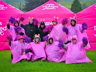 A group of 9 Tickled Pink volunteers at an outdoor event wearing rain ponchos and smiling as they wave pink and purple cheerleader pompoms. In the background are pink marquees with the Asda Tickled Pink logo on them.