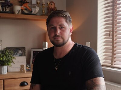A photo of Alex sitting at a table in a home. He has short brown hair, a beard, tattoos and is wearing a dark t-shirt.