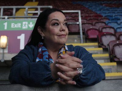 A photo of Lisa sitting in an empty football stadium. She has long dark hair and is wearing a blue jacket and a football scarf.