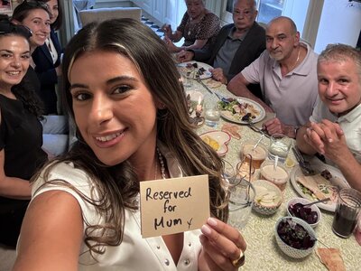 Evie is taking a selfie of the whole family around the dinner table. In one hand she's holding a hand written sign that says, 'Reserved for Mum x'