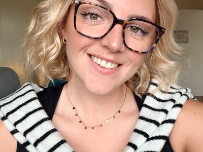 Selfie of a woman with blonde short hair and glasses. She is smiling at the camera wearing a black top with a black and white striped jumper tied around her shoulders.