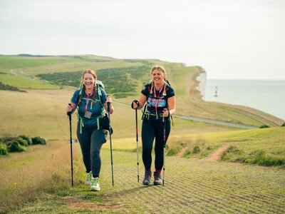Photo of 2 women with outdoor gear and walking poles taking part in the South Coast Ultra Challenge. In the background is countryside and chalk cliffs by the sea.