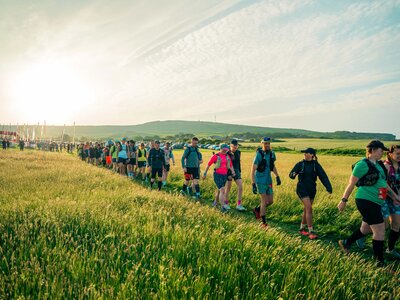 Photo of a long line of people walking through a sunny field as they begin the Isle of Wight Ultra Challenge. The start line and flags can be seen in the background.