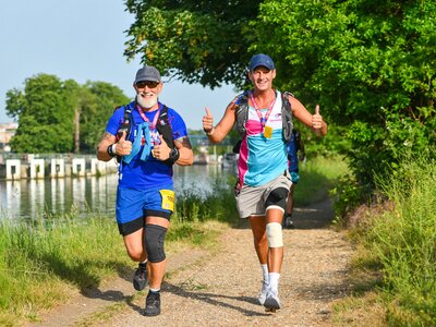 Photo of 2 men in running gear giving thumbs up as they run along a path by a river as part of the London to Brighton Ultra Challenge