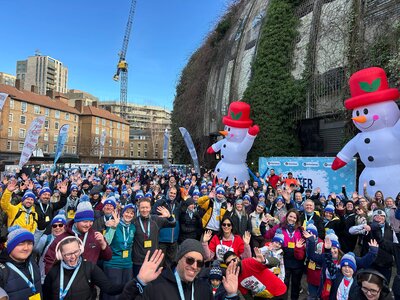 A photo of a large group of London Winter Walk participants, many of them wearing matching bobble hats, waving and smiling at the camera.