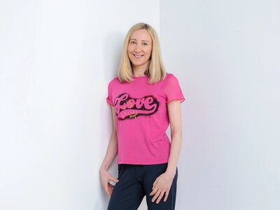 Christina has blonde hair. She's wearing a pink T-shirt with the word 'Love' on it with black leggings and pink high heels.